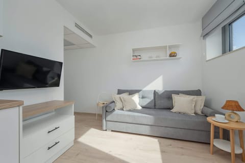 A bright living room with large windows allowing natural light in, featuring a sofa, coffee table, and a wall-mounted TV. The neutral tones and simple decor give the space a modern aesthetic.