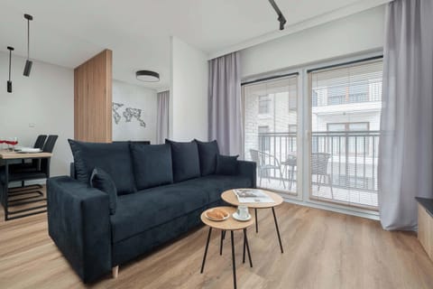 Bright living area with a dark grey sofa, light wooden flooring, and large windows covered with light curtains.