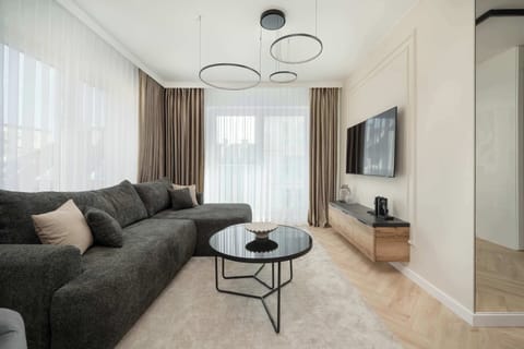 A modern living room with a dark grey sectional sofa, a round coffee table, and a large window letting in natural light. A chic chandelier hangs from the ceiling, adding an elegant touch to the room.