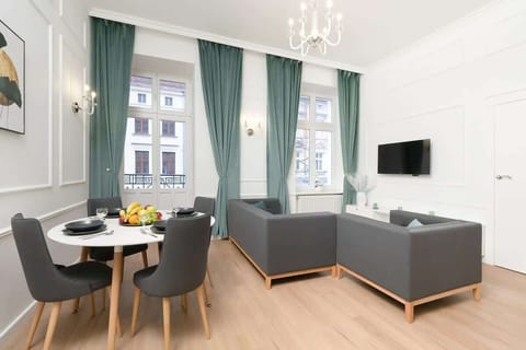 Bright living area with large windows, green curtains, a round dining table, gray sofa, and wall-mounted TV.

