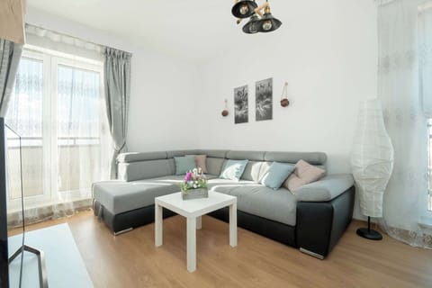 A spacious living room featuring a corner sofa in shades of gray, a white coffee table, and decorative lighting, creating a cozy atmosphere. Large windows let in plenty of natural light.