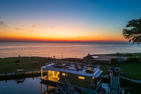 Aerial sunset view of the floating tiny home docked at Patapsco Marina on the Chesapeake Bay, the rooftop deck glowing above a quiet cove, gazebo, and open water.