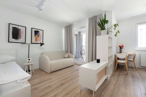A bright, open-plan studio features a minimalist white bed and a cozy cream sofa. The space is decorated with abstract wall art and light wood-effect flooring that leads toward a small dining area.