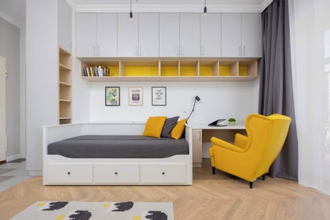 Discover a vibrant and functional studio, where a comfortable daybed with storage meets a bright yellow armchair and a dedicated workspace, all enhanced by ample shelving and cheerful yellow accents.