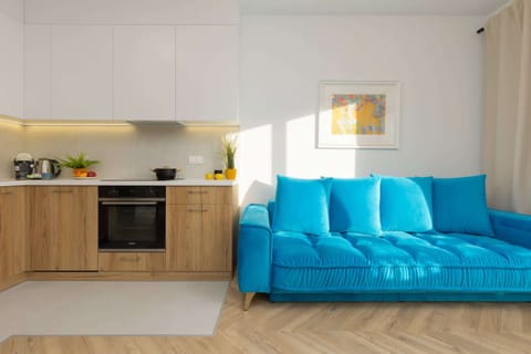 A modern living room with a bright blue sofa, a wooden floor, and an open kitchen with light wood cabinets. The space is well-lit with natural light.