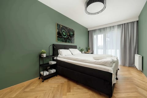 A cozy bedroom with a queen-sized bed featuring a white comforter. The room is decorated with green walls, light wood furniture, and stylish lighting fixtures.