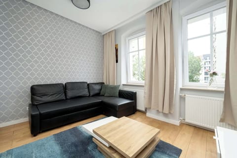 A bright living room featuring a black sofa, wooden floors, a small coffee table, and a TV set by the window.
