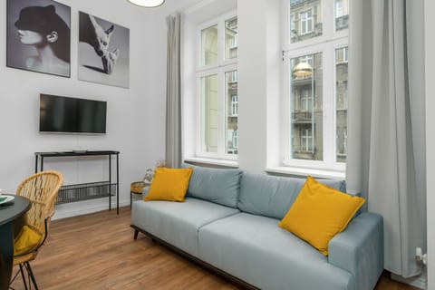 A modern living room with a grey sofa, yellow cushions, a small coffee table, and large windows allowing natural light.