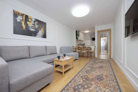 A cozy living room with a gray sofa, a rug, and a TV mounted on the wall. A wooden floor and soft lighting create a warm atmosphere.