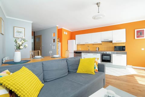A bright living room with light blue walls, a gray sectional sofa, yellow accent pillows, and an open kitchen with orange highlights in the background.