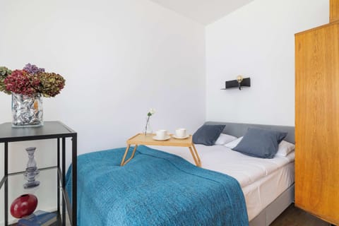 The bedroom has a cozy single bed with a blue blanket, a bedside table with a flower vase, and a wall-mounted light fixture. A tall wooden wardrobe stands nearby, providing ample storage.
