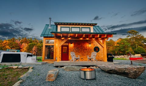 Evening light on the Cherry Cabin, with warm cedar siding, a red door, and two wooden chairs on the covered deck by the fire pit.