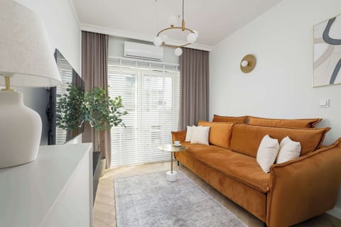 A modern living room with a large orange sofa and a coffee table, featuring access to a balcony through a glass door. The space is well-lit with natural light.