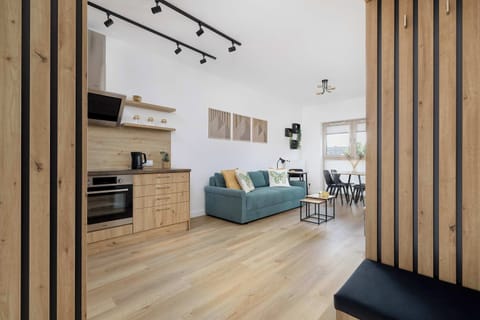 A modern, open-concept living space with light wooden flooring, a stylish kitchenette with wooden cabinets and a black range hood, a cozy teal sofa with decorative pillows, and a dining area by large windows creates a bright, functional interior.