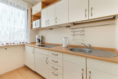 A modern kitchenette with light-colored cabinets and countertops, equipped with an electric kettle, coffee maker, and a functional sink, illuminated by natural light coming through a window with sheer curtains, creates a cozy and practical space for