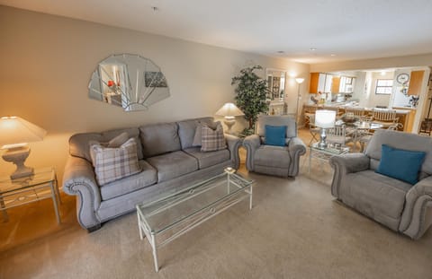 Gray living room seats with nailhead trim flank a glass-top coffee table on beige carpet, adjacent to the open dining area.