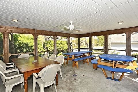 The airy screened porch features a large wooden dining table surrounded by white chairs and multiple blue picnic-style tables, perfect for leisurely meals outdoors.