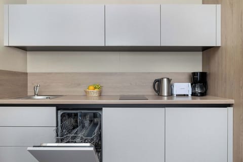 A modern kitchenette with white cabinets, an open dishwasher, kettle, toaster, coffee maker, and a basket of bananas – a functional space for everyday use.