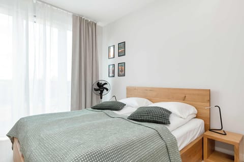 A bedroom area with a wooden bed frame, green bedspread, and white curtains letting in soft daylight.