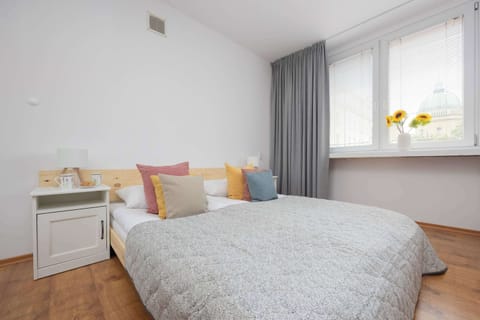 Bright studio area with a large bed, gray quilt, colorful pillows, white nightstand, wooden floor, and two windows with sheer curtains and a vase of yellow flowers on the windowsill.