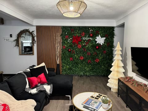Welcome to our festive living room! Relax on the cozy sectional sofa with holiday pillows & throws. Enjoy a stunning green accent wall with festive decor, a charming white Christmas tree, and a modern TV. A unique mirror and stylish coffee table complete
