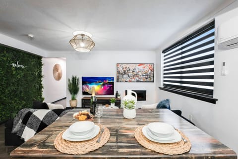 Chic open-concept space featuring a dining table set for four with woven placemats, leading to a cozy living area with a large TV, electric fireplace, and lush green accent wall. Modern lighting, air conditioning, and stylish blinds complete this inviting