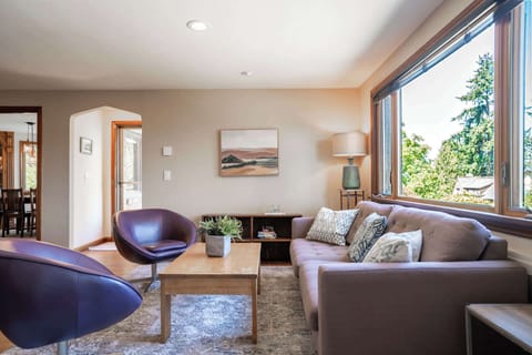 This bright, inviting living room offers a warm, relaxing experience with its large sunlit windows, cozy seating, and serene view of greenery outside.