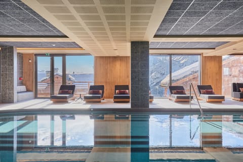 Pool deck with dark loungers and mountain rooftops beyond the glass — natural light fills the space throughout the day.