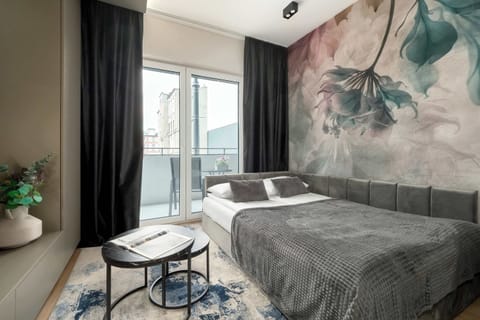 A cozy bedroom area with a large bed, grey bedding, and artistic wall mural in soft green and grey tones.

