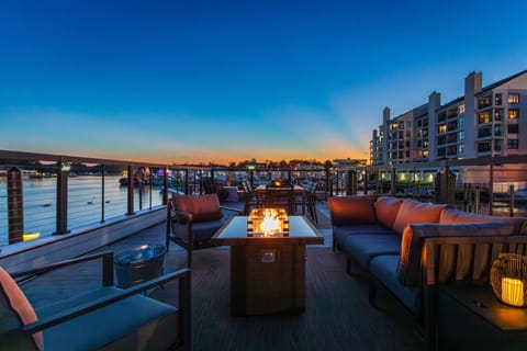 Rooftop deck glowing at blue hour on Rudee Inlet, with a lit fire pit, deep sectional, and full dining table overlooking the marina and the waterfront condos of Virginia Beach.