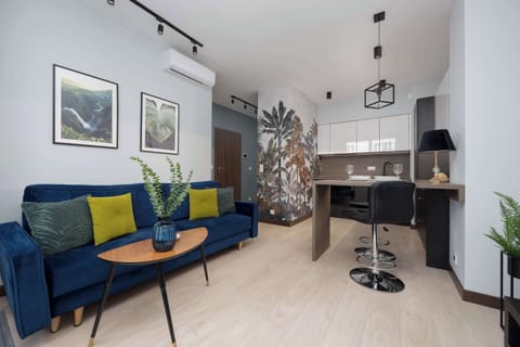 A cozy living room with a dark blue sofa accented by colorful cushions and modern artwork on the wall. The space is complemented by a small dining table and a compact kitchen area in the background.