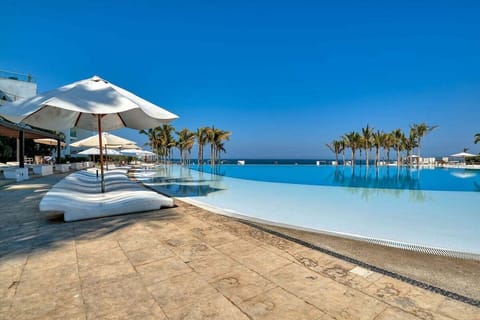 An expansive infinity pool blends seamlessly with the ocean under a clear blue sky, featuring white sun loungers and umbrellas for ultimate relaxation.