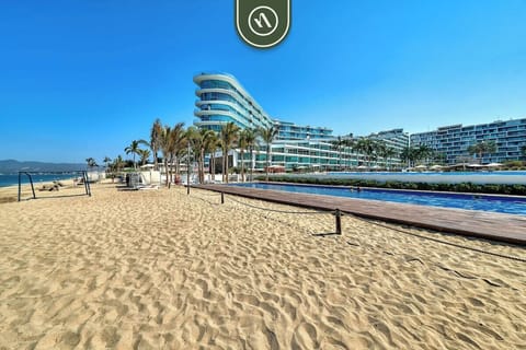A wide-angle view from the sandy shore looking toward the modern, multi-story resort. The long lap pool and swaying palm trees perfectly complement the sleek, glass-front design.