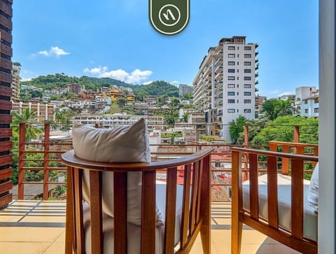Step out onto your private balcony featuring comfortable wooden chairs. Enjoy vibrant views of the surrounding city architecture and the lush, green hillside.