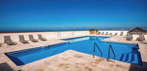 A beautiful rooftop pool area with lounge chairs lined up under a wide, sunny sky, offering expansive ocean views.