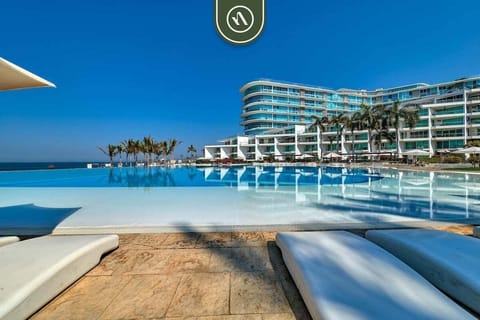 A stunning shot of the resort's expansive infinity pool reflecting the clear blue sky. Modern lounge chairs line the deck, while the architectural curves of the luxury apartment complex rise in the background.