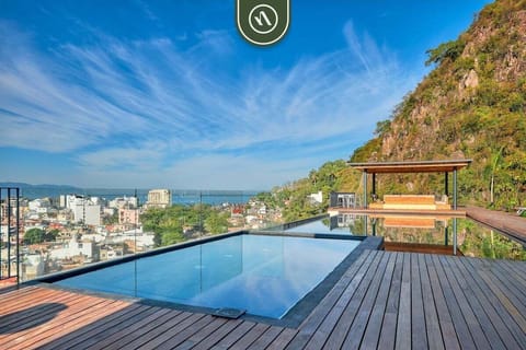 Stunning rooftop pool surrounded by wooden decking, offering views of the city, ocean, and lush mountainside.