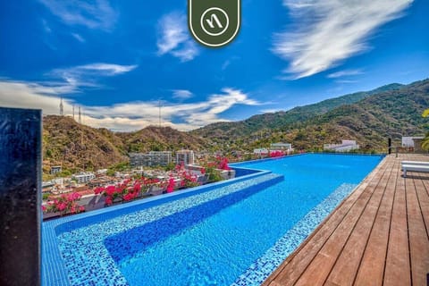 Beautiful rooftop infinity pool with a wooden deck, overlooking lush mountains and a bright blue sky.