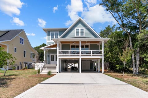 Elevated coastal home, wide driveway, carport, stairs, front porch, railings, glass doors, windows, trees, neighboring house, blue sky clouds.