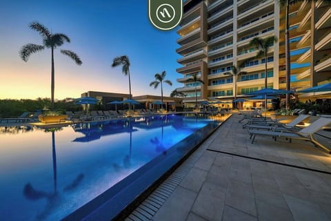 Relax by the large ground-floor swimming pool illuminated at twilight, surrounded by palm trees and lounge chairs.