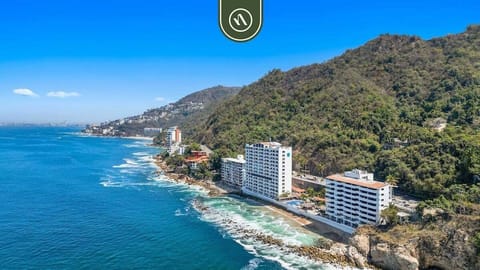 aerial view of a multi-story white hotel or residential building complex situated along a rocky coastline, featuring swimming pools, sandy patches, and a backdrop of lush green mountain slopes meeting the open ocean.