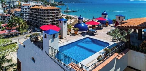 High-angle shot of a rooftop swimming pool complex featuring distinctive blue domes and red patio umbrellas overlooking the ocean.
