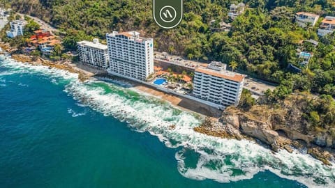 High-angle drone perspective of a white multi-story resort built on a rocky shoreline cove.