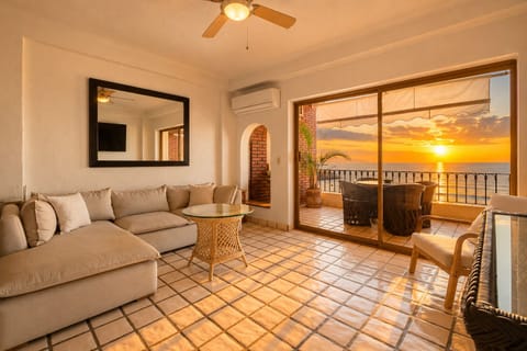 Bright living room opening directly to a furnished ocean-view balcony, with sunset views over