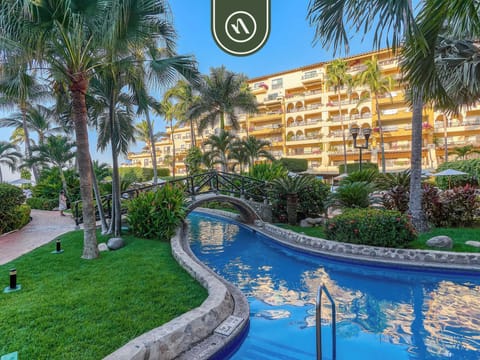 Scenic, river-style swimming pool lined with lush green grass and palm trees, featuring a charming stone footbridge.