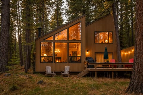 Modern cabin nestled among towering pines, featuring expansive windows that glow warmly at dusk, a welcoming deck with BBQ and seating, and tranquil forested surroundings.