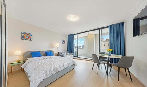 Bright studio-style bedroom featuring a comfortable queen bed, bedside tables with lamps, dining table set, wall-mounted TV, and sliding doors opening to a private balcony.