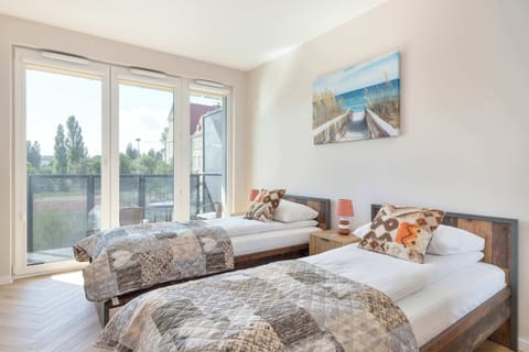 Bright studio featuring two single beds with patterned bedding, a large window, and a scenic wall painting.