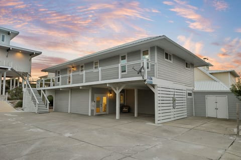 Coastal-style vacation home features stilted construction and ample parking, with sunset-painted skies above the neighboring beach community.