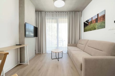 A cozy living room with a beige sofa bed and a TV mounted on the wall. Soft curtains and a painting create a warm, welcoming atmosphere.
